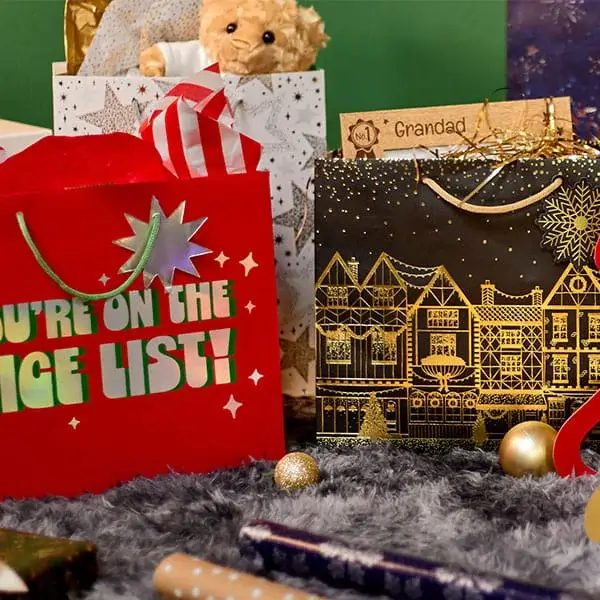 Festive Christmas gift bags filled with gifts, cards and a teddy bear, displayed on a soft grey rug with gold baubles and wrapped presents.